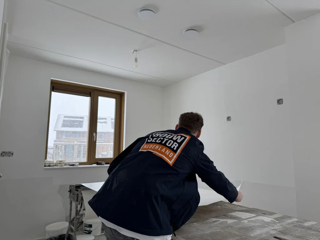 Behanger in werkjas van Bouwsector Nederland zet een baan tegen de wand van een nieuwbouwkamer terwijl het buiten sneeuwt