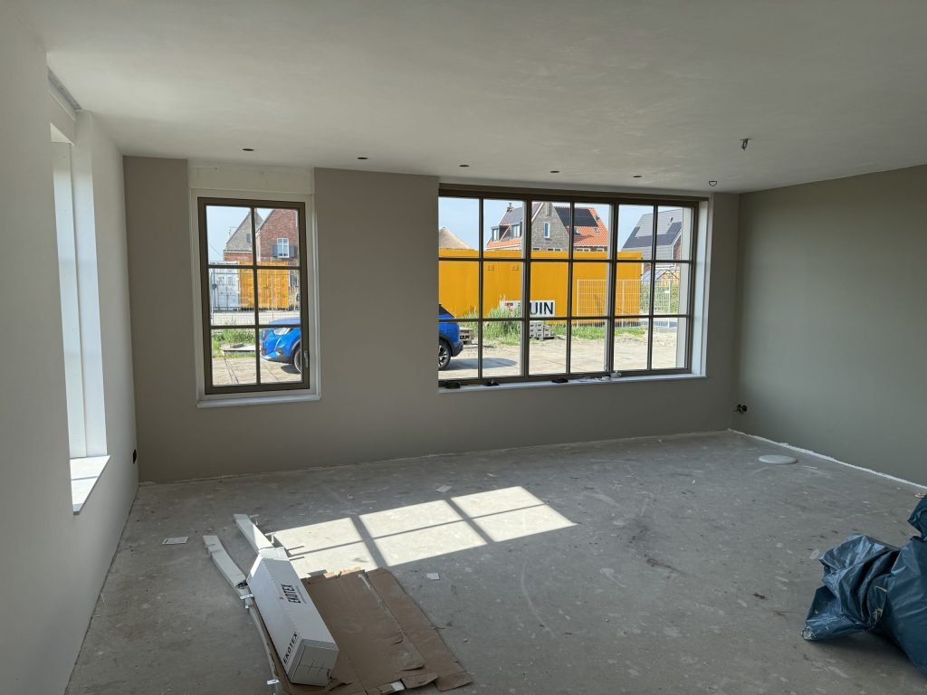 Woonkamer van een nieuwbouwwoning met grijsgroene wanden, grote roedenramen en een betonvloer