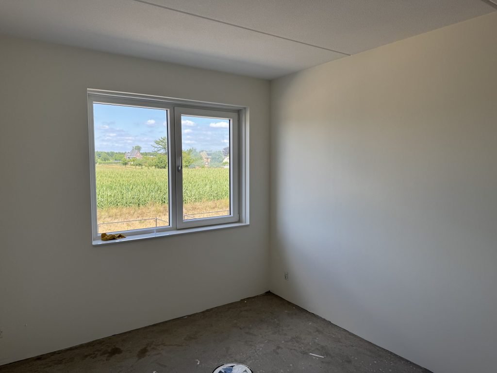 Lege slaapkamer in een nieuwbouwwoning met strak afgewerkte witte wanden en een raam met uitzicht op een maisveld
